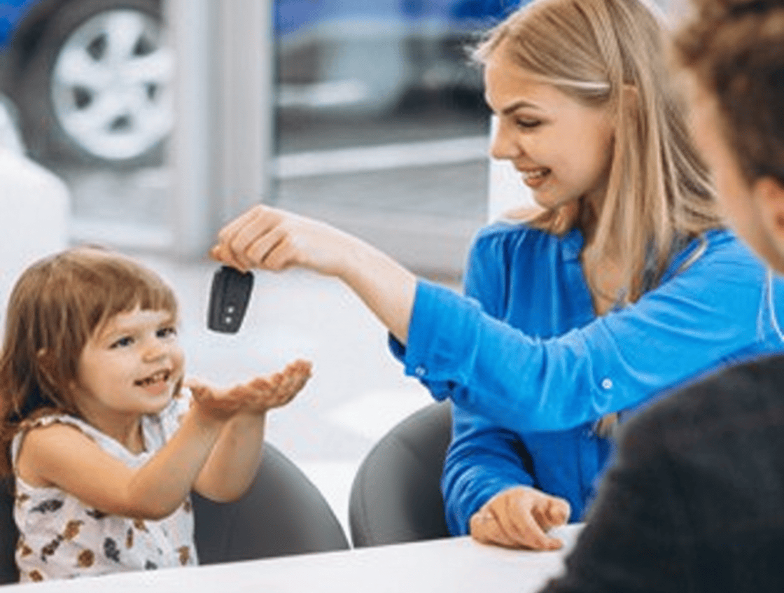 Une femme blonde et sa fille de 4 ans sont assises dans une concession automobile. La femme donne les clés de sa nouvelle voiture à sa fille en souriant ; la petite fille tend les mains pour recevoir le cadeau.