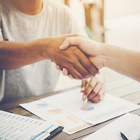 Photo de deux personnes se serrant la main au dessus d'une table sur laquelle des documents sont dispersés.